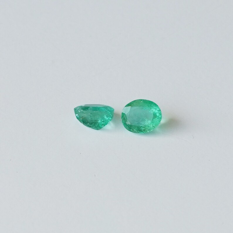 May include: Two emerald green gemstones, one slightly larger than the other, are displayed on a white surface.
