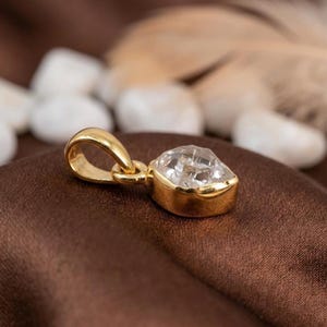 K&ouml;nnte beinhalten: Ein goldener Anh&auml;nger mit einem klaren, facettierten Edelstein. Der Anh&auml;nger hat eine abgerundete Form mit einer goldenen &Ouml;se f&uuml;r eine Kette. Der Edelstein ist in einer goldenen L&uuml;nette gefasst, auf braunem Untergrund.