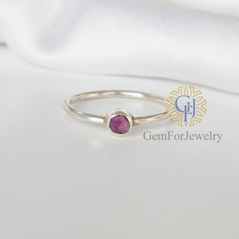 May include: A delicate silver ring with a small, faceted pink gemstone set in a bezel setting. GemForJewelry is printed below the ring.
