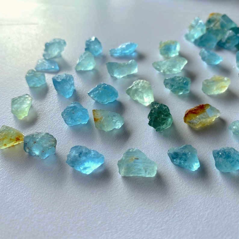 Pode incluir: Uma cole&ccedil;&atilde;o de gemas de &aacute;gua-marinha em v&aacute;rios tons de azul, verde e amarelo. Os cristais brutos e naturais s&atilde;o exibidos em uma superf&iacute;cie branca.