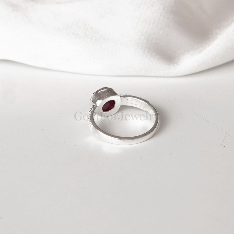 Peut inclure: Une bague en argent avec une pierre pr&eacute;cieuse rouge sertie au centre. La bague a une bande de petites pierres claires.