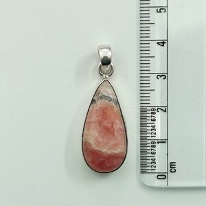 May include: A teardrop-shaped pendant with a pink and white stone set in a silver-colored metal. The pendant is approximately 1.5 inches long. The stone has a layered appearance with bands of color. A ruler with centimeter markings is in the background.