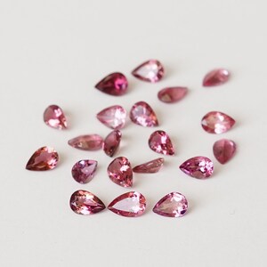 May include: A collection of pink pear-shaped gemstones, scattered on a white surface.