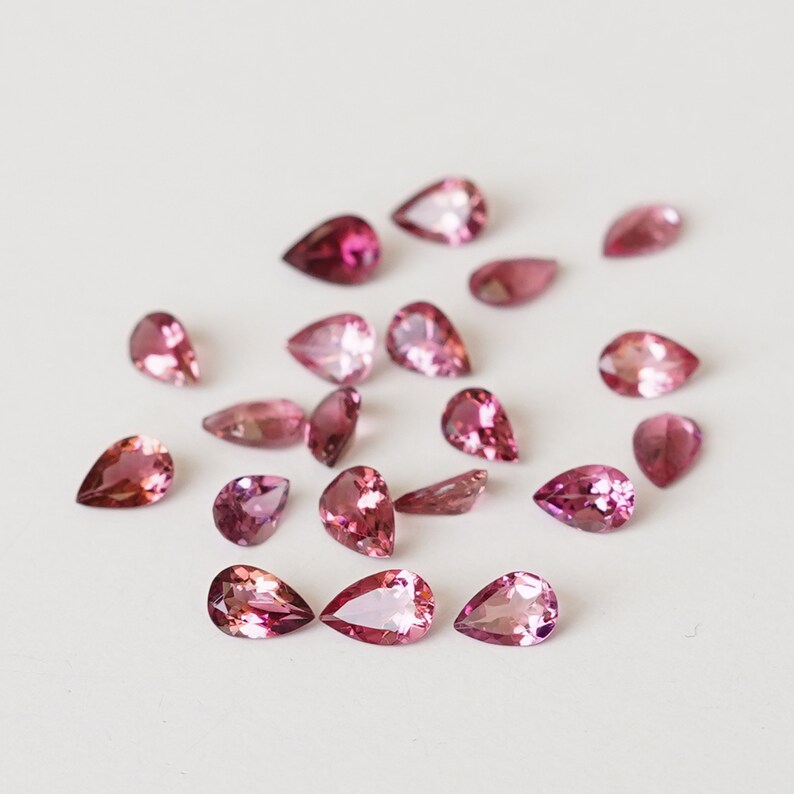 May include: A collection of pink pear-shaped gemstones, scattered on a white surface.