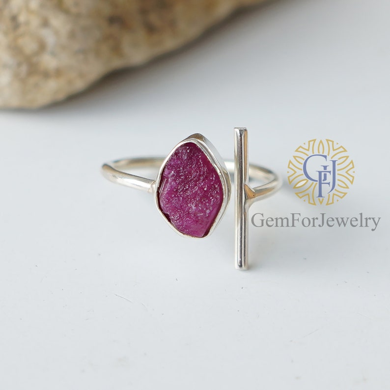 K&ouml;nnte beinhalten: Ein silberner Ring mit einem rohen rosafarbenen Rubin-Edelstein und einem silbernen Balkenakzent. Der Ring liegt auf einem wei&szlig;en Hintergrund. 'GemForJewelry' ist auf dem Hintergrund gedruckt.