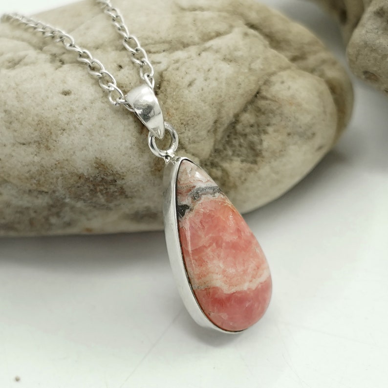 May include: A teardrop-shaped pendant with a pink and white stone, set in a silver-colored metal frame. The pendant hangs from a delicate silver chain. The stone has a layered appearance with black and white inclusions.
