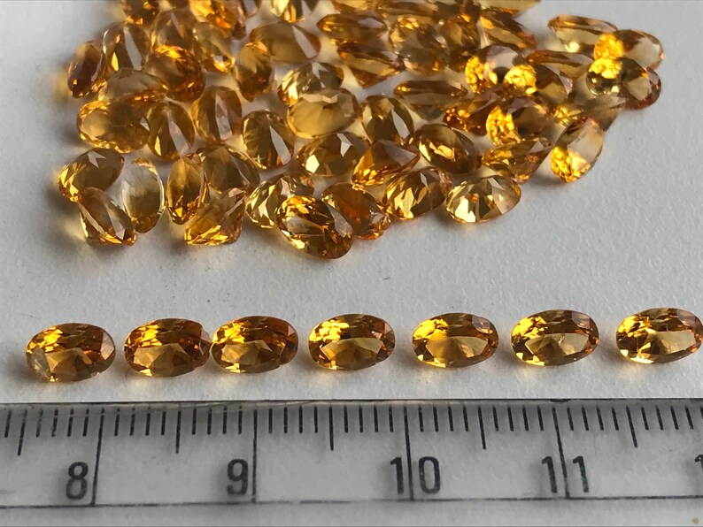 May include: A close-up of a group of oval-shaped, faceted, golden-brown gemstones. The gemstones are arranged in a pile on a white surface. A ruler is placed below the gemstones, showing measurements in inches.