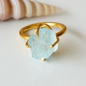 May include: Gold ring featuring a light blue aquamarine gemstone. The gemstone is a rough, natural shape.