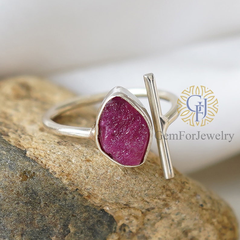 K&ouml;nnte beinhalten: Ein silberner Ring mit einem rohen rosafarbenen Rubin-Edelstein und einem silbernen Balkenakzent. Der Ring liegt auf einem steinernen Hintergrund. Der Text 'GemForJewelry' ist auf dem Hintergrund gedruckt.