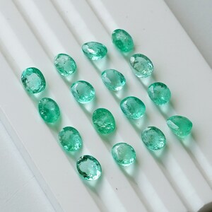 May include: A collection of 15 oval-shaped, faceted, green gemstones. The gemstones are arranged in rows on a white surface.