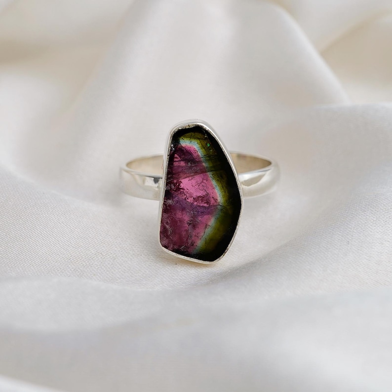 K&ouml;nnte beinhalten: Ein Silberring mit einem gro&szlig;en, unregelm&auml;&szlig;ig geformten Wassermelonen-Turmalin-Edelstein. Der Edelstein zeigt einen lebendigen Farbverlauf von Rosa, Gr&uuml;n und Schwarz. Der Ring ist vor einem wei&szlig;en, seidigen Hintergrund platziert.