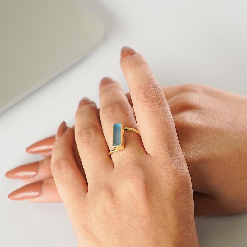 May include: A gold ring featuring a single, rectangular, light blue stone set vertically. The band is thin and metallic, showcasing a polished gold finish that contrasts with the cool, translucent tone of the elongated blue gemstone centerpiece.
