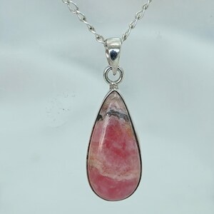 May include: A teardrop-shaped pendant with a pink and white stone, set in a silver-colored metal frame. The pendant hangs from a delicate silver chain. The stone has a marbled appearance with some black inclusions.