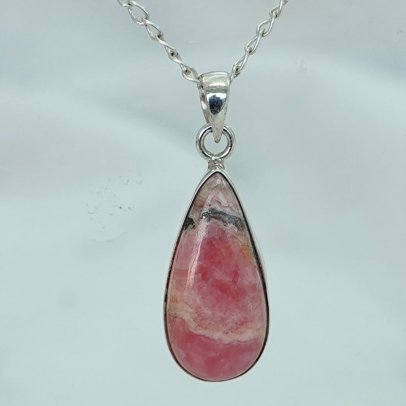 May include: A teardrop-shaped pendant with a pink and white stone, set in a silver-colored metal frame. The pendant hangs from a delicate silver chain. The stone has a marbled appearance with some black inclusions.