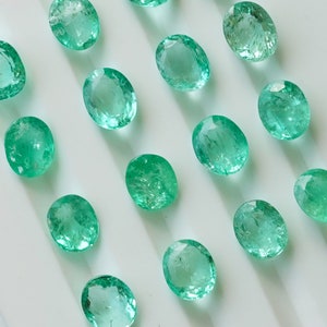 May include: A collection of oval-shaped, green gemstones. The gemstones are arranged in rows on a white surface.