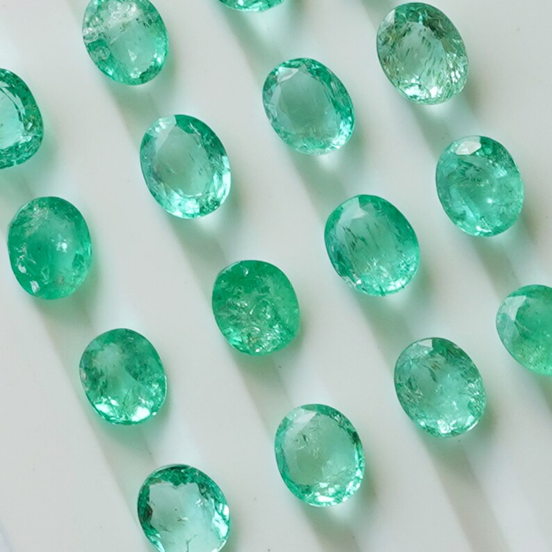 May include: A collection of oval-shaped, green gemstones. The gemstones are arranged in rows on a white surface.