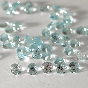 K&ouml;nnte beinhalten: Eine Sammlung von hellblauen Edelsteinen, einige sind facettiert und andere rund, verstreut auf einer wei&szlig;en Oberfl&auml;che.