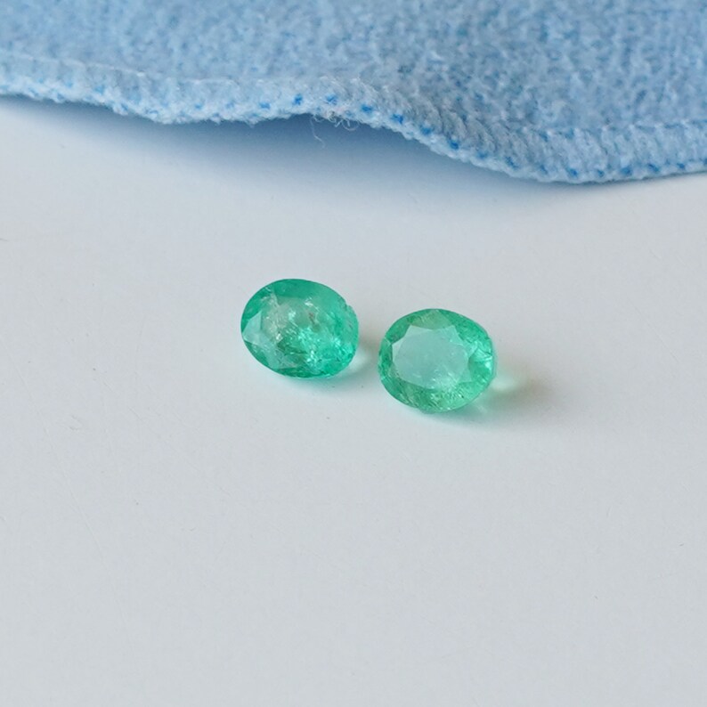 May include: Two oval-shaped emerald green gemstones on a white surface.