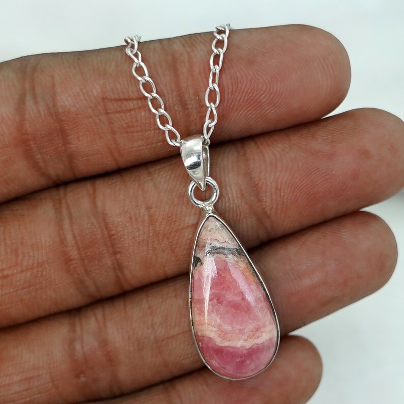 May include: A silver necklace featuring a teardrop-shaped pendant. The pendant has a pink and white stone with a silver bezel. The chain is made of small, linked silver pieces. The necklace is displayed against a neutral background.