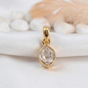 K&ouml;nnte beinhalten: Ein goldener Anh&auml;nger mit einem klaren, facettierten Edelstein in Diamantenform. Der Anh&auml;nger hat oben eine kleine Schlaufe f&uuml;r eine Kette. Der Hintergrund ist ein weicher, wei&szlig;er Stoff.