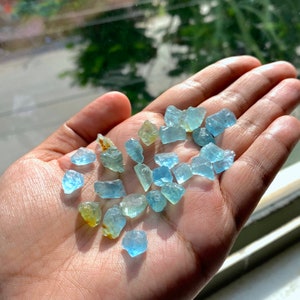 Pode incluir: Uma cole&ccedil;&atilde;o de pequenas gemas de &aacute;gua-marinha &aacute;speras, de cor azul claro, em v&aacute;rios formatos e tamanhos. As gemas est&atilde;o espalhadas na m&atilde;o de uma pessoa.