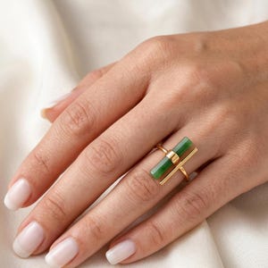 May include: A gold ring featuring a rectangular, vibrant green stone. The ring has a minimalist design with a horizontal bar and a vertical bar, creating a modern and elegant look. The ring is shown on a hand with well-manicured nails.