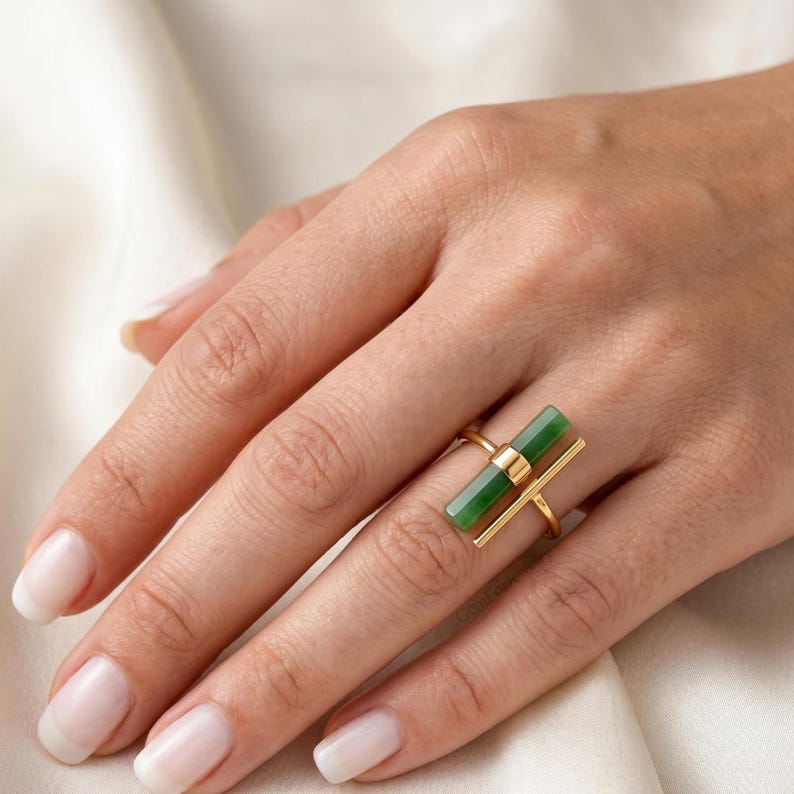 May include: A gold ring featuring a rectangular, vibrant green stone. The ring has a minimalist design with a horizontal bar and a vertical bar, creating a modern and elegant look. The ring is shown on a hand with well-manicured nails.