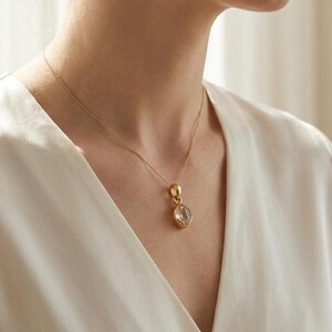 K&ouml;nnte beinhalten: Eine goldfarbene Halskette mit einem klaren, facettierten Anh&auml;nger. Der Anh&auml;nger hat eine Diamantenform und ist in eine goldene L&uuml;nette gefasst. Die Halskette wird &uuml;ber einem wei&szlig;en Oberteil getragen.