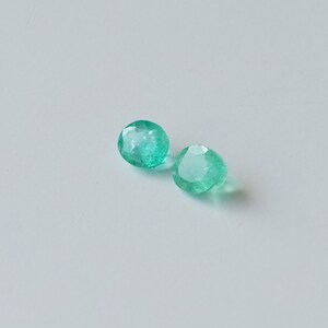 May include: Two round, faceted, green emerald gemstones on a white background.