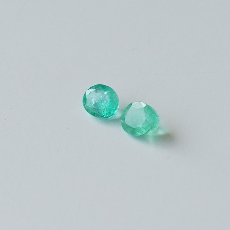 May include: Two round, faceted, green emerald gemstones on a white background.