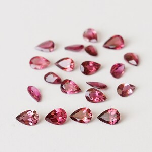May include: A collection of pink pear-shaped gemstones, scattered on a white surface.