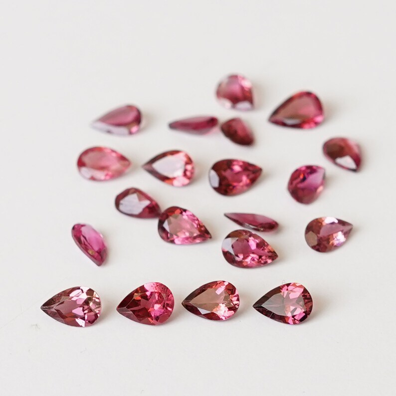 May include: A collection of pink pear-shaped gemstones, scattered on a white surface.