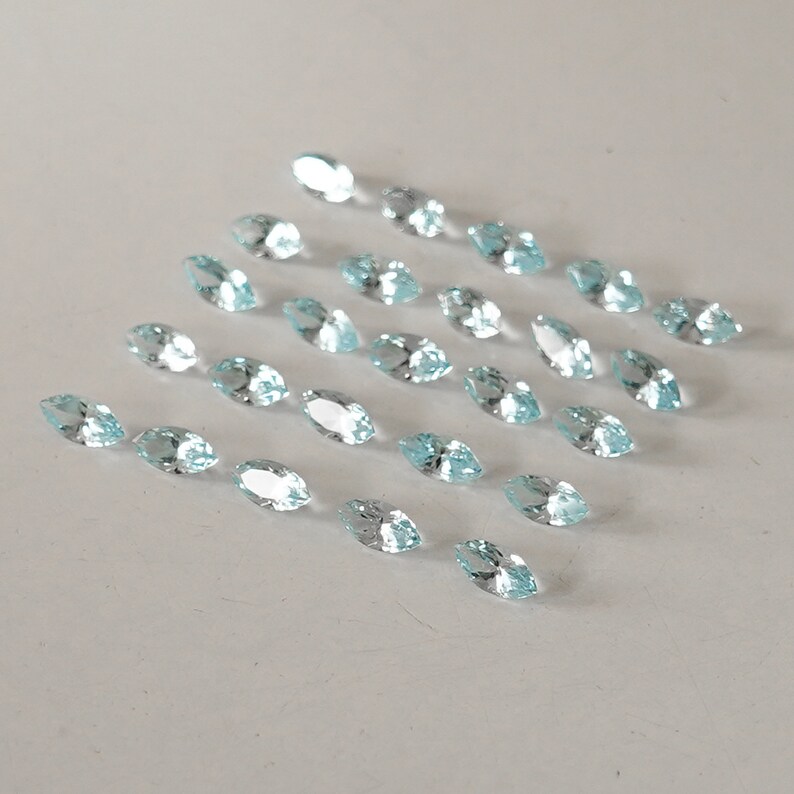 May include: A collection of light blue marquise cut gemstones arranged in a triangular pattern on a white surface.