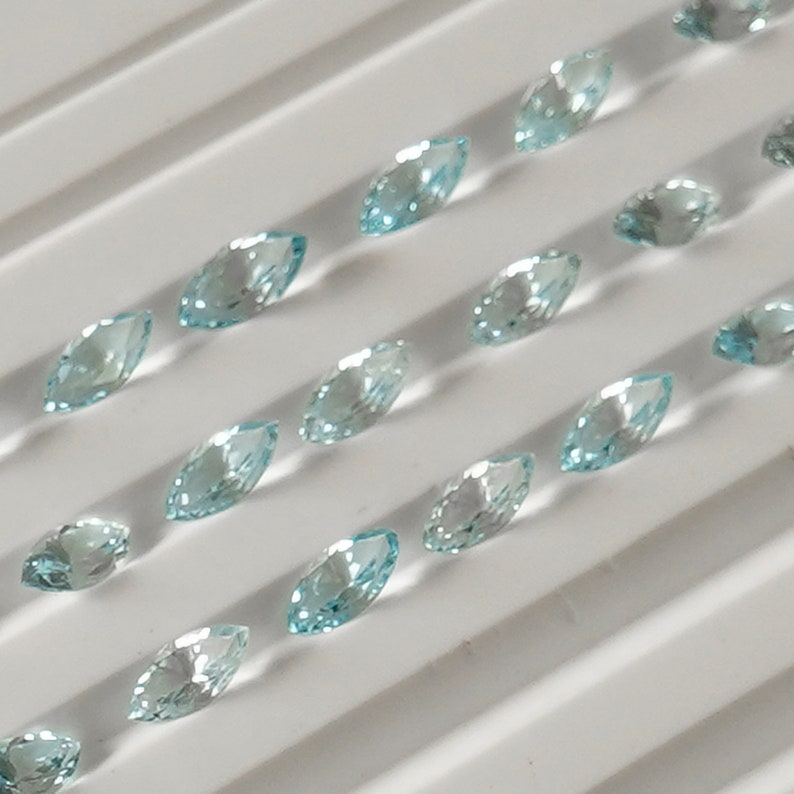 May include: A collection of light blue marquise-shaped gemstones arranged in rows on a white background.