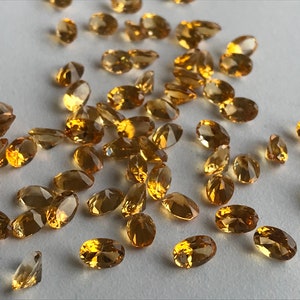 May include: A collection of oval-shaped, faceted, golden citrine gemstones. The citrines are scattered on a white surface.