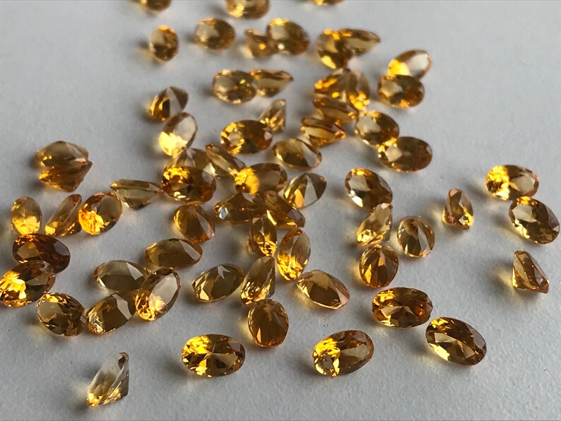 May include: A collection of oval-shaped, faceted, golden citrine gemstones. The citrines are scattered on a white surface.