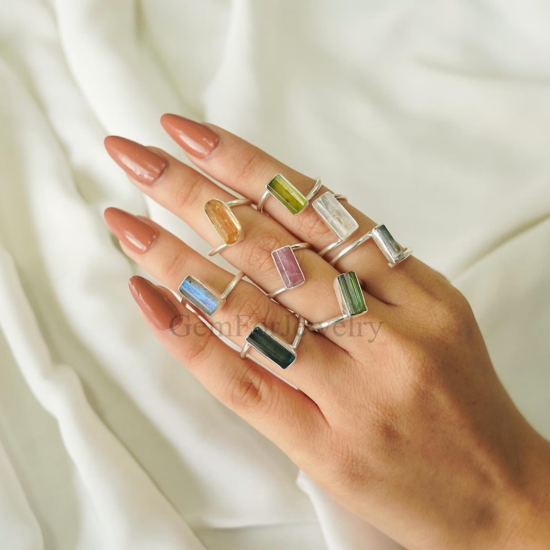 May include: A collection of seven silver rings, each featuring a single rectangular, baguette-cut gemstone in various colors including yellow, green, pink, blue, and clear, worn on multiple fingers to showcase the diverse mineral hues.