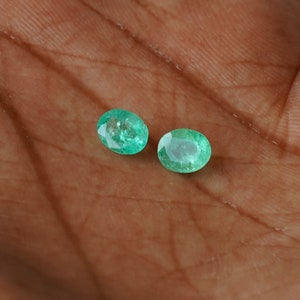 May include: Two oval-shaped emerald gemstones, a vibrant green color, are displayed on a brown surface.