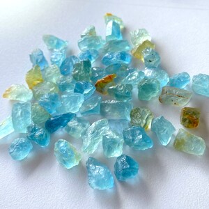 Pode incluir: Uma cole&ccedil;&atilde;o de gemas de &aacute;gua-marinha bruta em v&aacute;rios tons de azul e verde. As pedras t&ecirc;m uma forma irregular e uma apar&ecirc;ncia transl&uacute;cida.