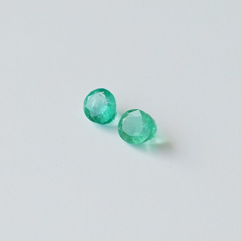 May include: Two round, faceted, green emerald gemstones.