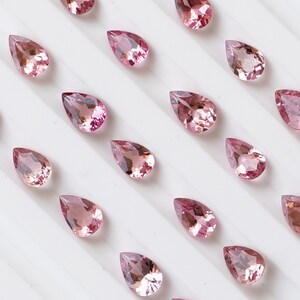 May include: A collection of pink pear-shaped gemstones arranged on a white surface. The gemstones are all different sizes and have a subtle sparkle.