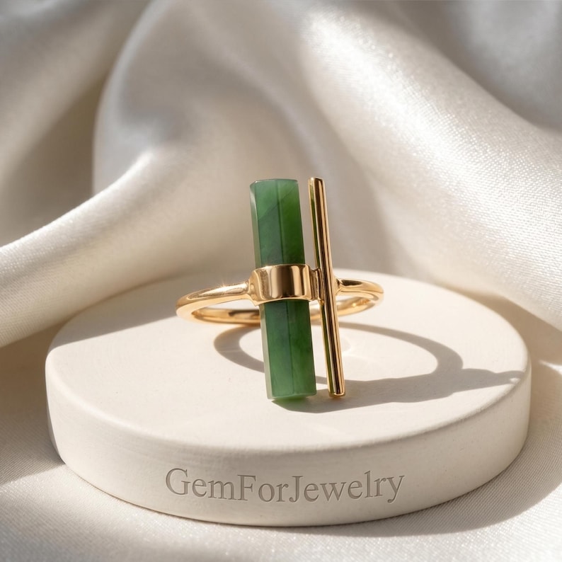 May include: A gold ring featuring a vertical green jade stone and a vertical gold bar. The ring is set on a white circular display with the text "GemForJewelry" at the base. The background is a white, silky fabric.