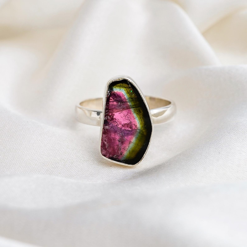 K&ouml;nnte beinhalten: Ein Silberring mit einem gro&szlig;en, unregelm&auml;&szlig;ig geformten Wassermelonen-Turmalin-Edelstein. Der Edelstein zeigt eine lebendige Mischung aus rosa, gr&uuml;n und schwarzen Farben. Der Ring ist vor einem wei&szlig;en, seidigen Hintergrund platziert.
