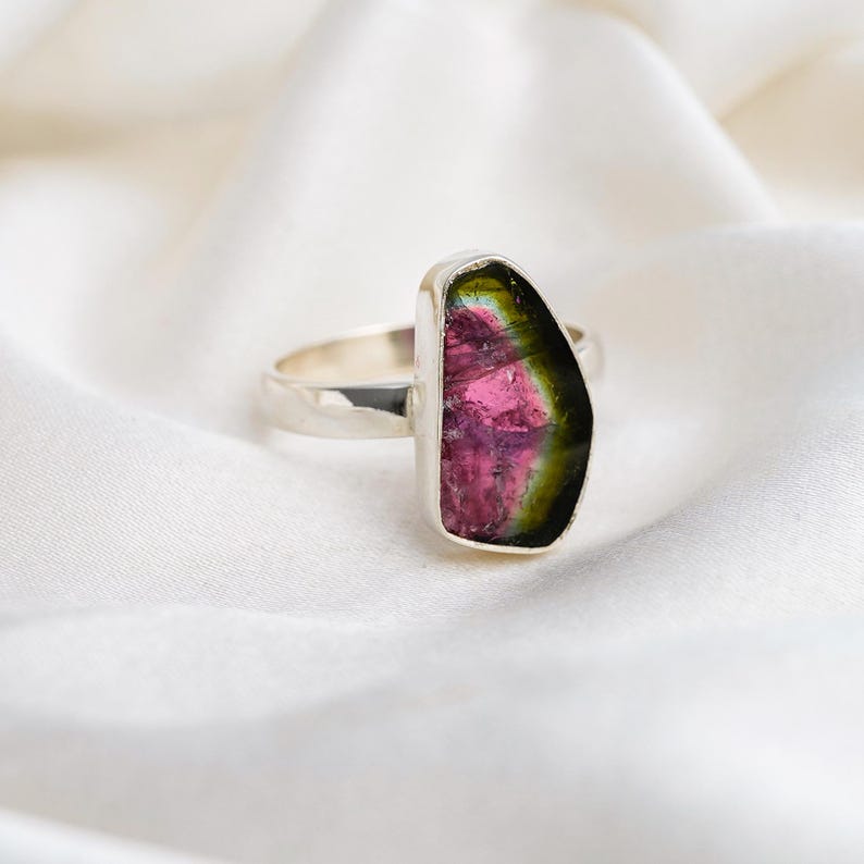 K&ouml;nnte beinhalten: Ein Silberring mit einem gro&szlig;en, roh geschnittenen Turmalin-Edelstein. Der Stein zeigt einen lebendigen Farbverlauf mit rosa, gr&uuml;nen und gelben Farbt&ouml;nen. Der Ring ist vor einem wei&szlig;en, strukturierten Hintergrund platziert.
