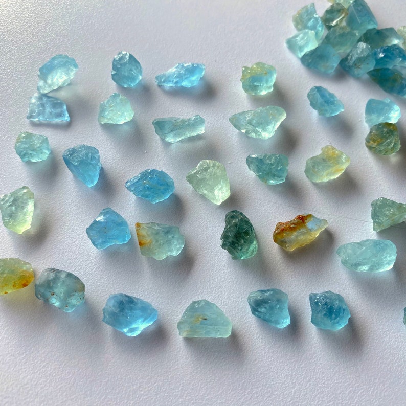 Pode incluir: Uma cole&ccedil;&atilde;o de cristais de &aacute;gua-marinha brutos em v&aacute;rios tons de azul e verde. Os cristais s&atilde;o &aacute;speros e t&ecirc;m um aspecto natural n&atilde;o polido.