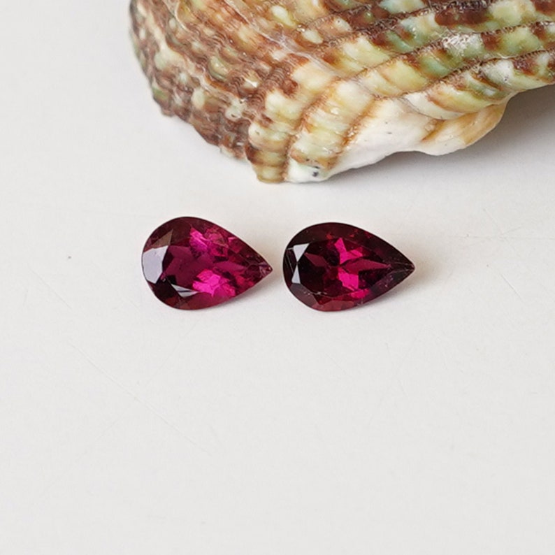 May include: Two pear-shaped gemstones, each a deep, rich red color. The gemstones are set against a white background.