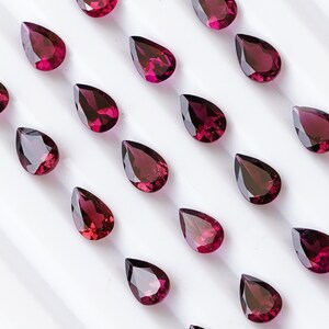 May include: A collection of pear-shaped, deep red gemstones arranged in rows on a white surface. The gemstones are all the same size and shape, and they are arranged in a symmetrical pattern.
