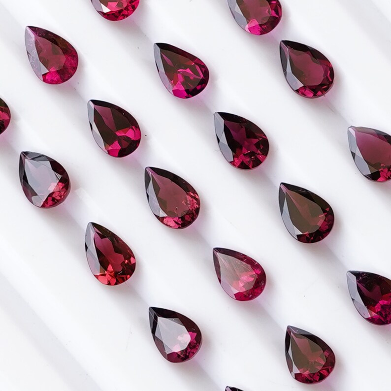 May include: A collection of pear-shaped, deep red gemstones arranged in rows on a white surface. The gemstones are all the same size and shape, and they are arranged in a symmetrical pattern.