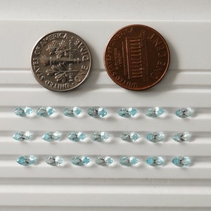 May include: Twenty-four marquise-shaped gemstones arranged in three rows of eight. The gemstones are a light blue color and are displayed on a white surface. The gemstones are approximately the size of a dime.