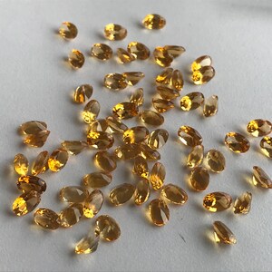 May include: A collection of oval-shaped, faceted, golden-yellow gemstones. The gemstones are scattered on a white surface.